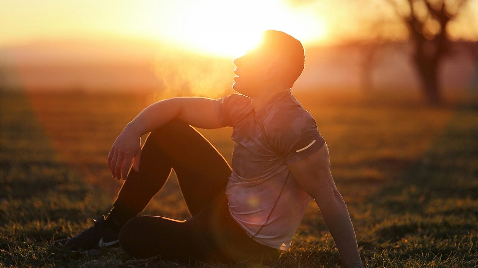 Method Muscular man sits in rural field at sunset, exhaling visible breath.